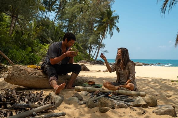 In a tropical setting, Rachal McAdams as Linda Liddle playfully engages with Dylan O'Brien as Bradley Preston, who enjoys a coconut while sitting by a small fire on the sandy beach. Lush greenery and the ocean are visible in the background.