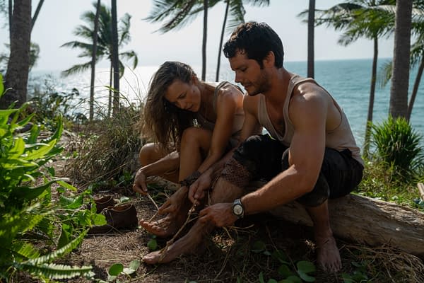 In a scene from 20th Century Studios' 'SEND HELP,' characters Linda Liddle and Bradley Preston work together on a beach, tying makeshift bindings on their feet. The backdrop features a serene ocean view and palm trees.