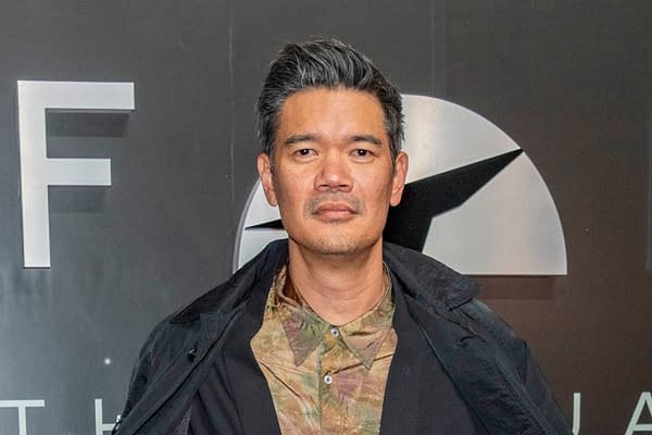 A man with short, grayish hair stands against a dark background with a prominent logo. He wears a patterned shirt and a black jacket, looking directly at the camera.