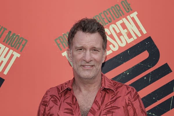 Thomas Jane attends a film premiere in Beverly Hills, wearing a red floral shirt against a vibrant backdrop featuring promotional text for a film. His expression is friendly and approachable.