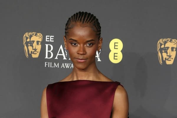 Letitia Wright poses confidently at the BAFTA Film Awards, wearing a stylish maroon dress and statement earrings, with the BAFTA logo visible in the background.