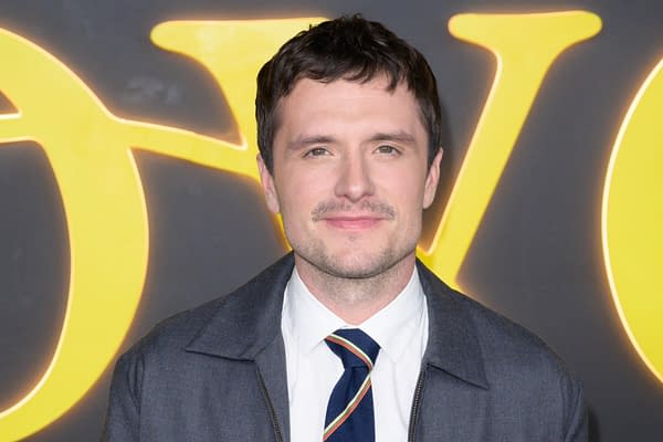 A man with short, dark hair and a light beard smiles at the camera. He is wearing a white shirt with a navy tie and a dark jacket, standing in front of a large yellow logo in a blurred backdrop.