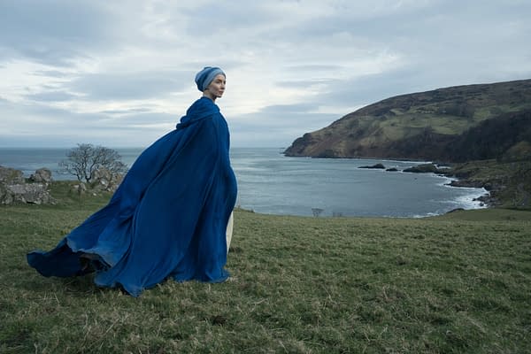 A woman in a flowing blue cloak stands on a grassy hillside overlooking a tranquil seascape with rocky formations and a distant coastline. The scene conveys a sense of solitude and contemplation.