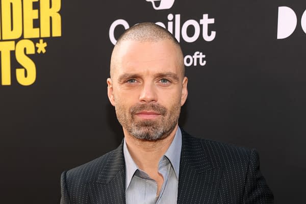 A man with a close-cropped hairstyle and short beard is smiling slightly, wearing a pinstriped suit over a light shirt. The background is dark with a logo and text, suggesting a promotional event or premiere.