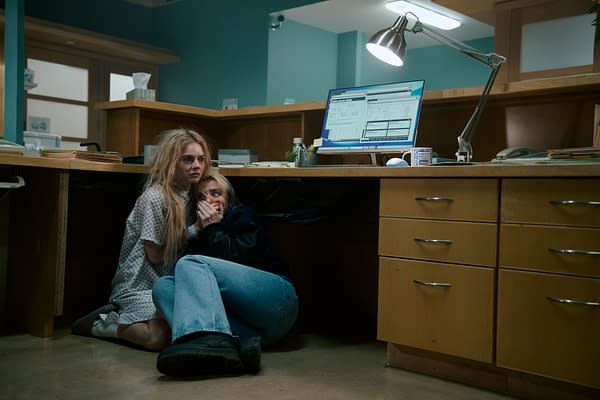 Two women crouch in fear under a desk in a hospital-like setting, one wearing a hospital gown and the other in casual clothing. The scene is tense, suggesting a moment of suspense from the film 'READY OR NOT 2: HERE I COME.'