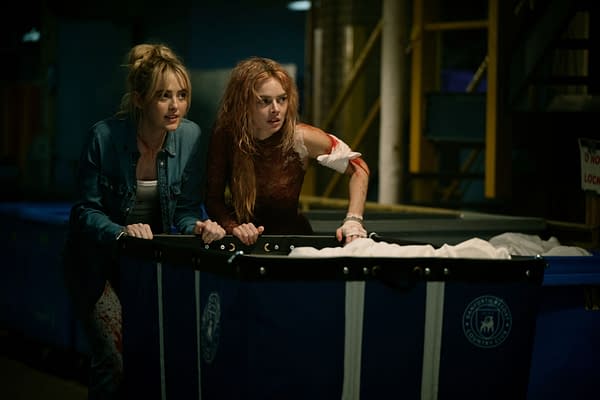 Two women, Kathryn Newton and Samara Weaving, looking nervously over the edge of a large blue container in a dimly lit industrial setting. One woman has blood on her arm and both appear tense, suggesting a moment of suspense from the film 'READY OR NOT 2: HERE I COME.'