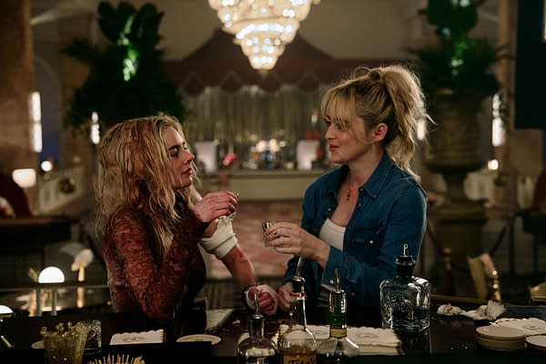 Samara Weaving and Kathryn Newton clink shot glasses at a bar in a scene from 'READY OR NOT 2: HERE I COME,' showcasing a lively atmosphere with ornate lighting and a stylish backdrop.