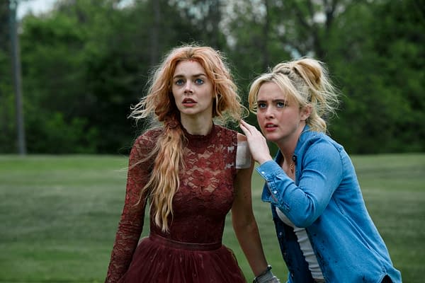 A scene from the film 'Ready or Not 2: Here I Come', featuring two women: one with long, wavy red hair wearing a burgundy lace dress, and the other with blond hair in a ponytail, dressed in a blue denim shirt. They appear anxious and are looking off-screen, set against a green outdoor background.