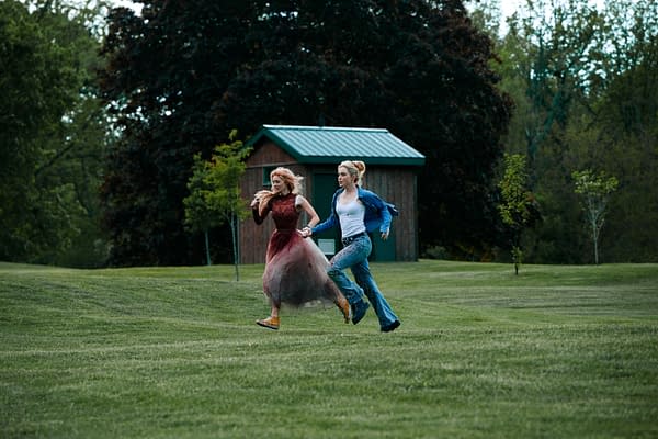 Samara Weaving and Kathryn Newton running together through a grassy field, with a wooden shed in the background. Weaving wears a flowy, burgundy dress while Newton is dressed in a casual blue outfit.