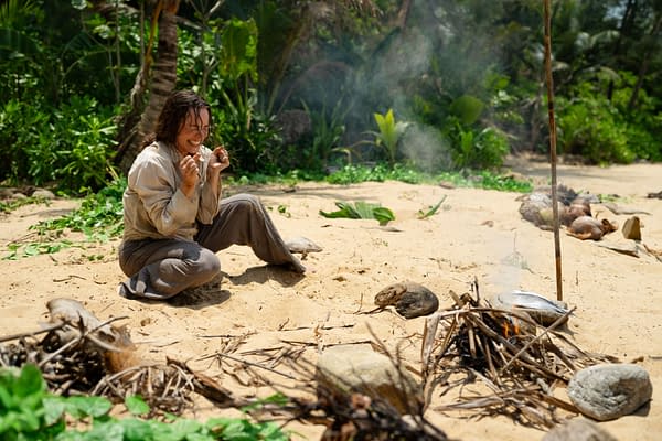 A scene from 20th Century Studios' 'SEND HELP' featuring Rachel McAdams as Linda Liddle, expressing fear or distress while sitting on a sandy beach surrounded by greenery and smoke rising from a small fire.