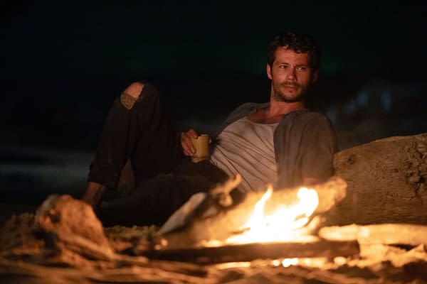A scene from the film 'SEND HELP' featuring Dylan O'Brien as Bradley Preston, sitting casually near a campfire while holding a drink, with a dark background highlighting the flames.