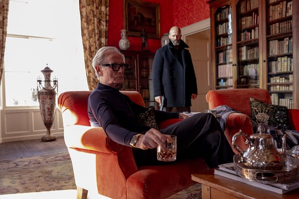 A scene from the film 'Untitled Statham Project' featuring an older man sitting on an orange couch, holding a drink, while another man stands in the background, dressed in a dark coat and holding a handgun. The setting includes rich red walls, bookshelves, and decorative objects.