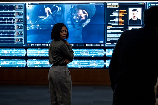 A woman with curly hair stands with a thoughtful expression in front of a large screen displaying digital data and an image of a man wearing a beanie, indicating a high-tech analysis setting.