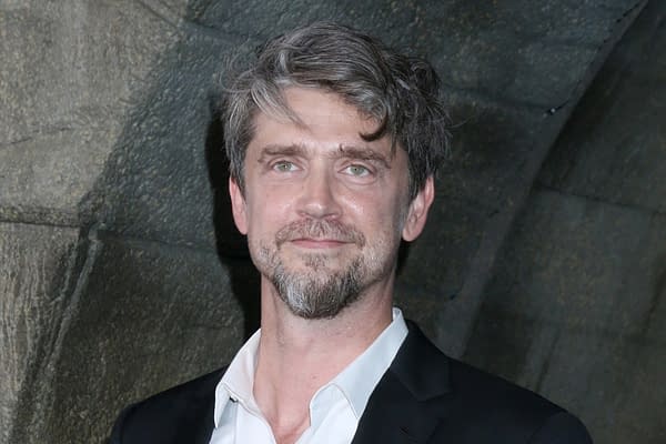 A man with wavy gray hair and a beard smiles at the camera, wearing a black suit and white shirt. He stands against a textured stone background.