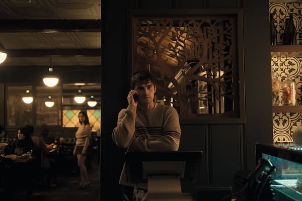 A scene from 'Obsession' features a young man in a beige sweater talking on the phone while standing beside a register. In the background, a woman in a light outfit appears to be walking through a dimly lit restaurant, surrounded by patrons.