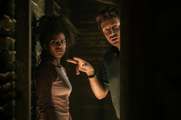 A tense scene in a dimly lit location featuring a woman with curly hair looking worried while a man gestures with his hand as if instructing her. The setting has a dark, eerie atmosphere with wooden walls.