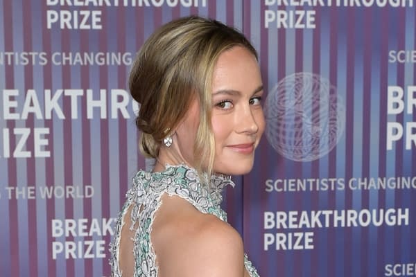 Brie Larson poses elegantly, wearing a detailed dress with floral designs, at the Breakthrough Prize event, featuring a backdrop with the event's name and slogan.