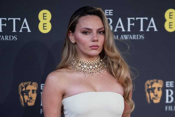 A woman with long, wavy hair wearing a glamorous pearl necklace and a strapless white gown poses confidently at the BAFTA Film Awards backdrop.