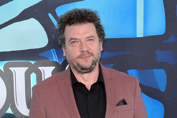 A man with curly hair and a beard is wearing a stylish jacket with a black shirt underneath, standing against a colorful blue backdrop. The expression is friendly and approachable.