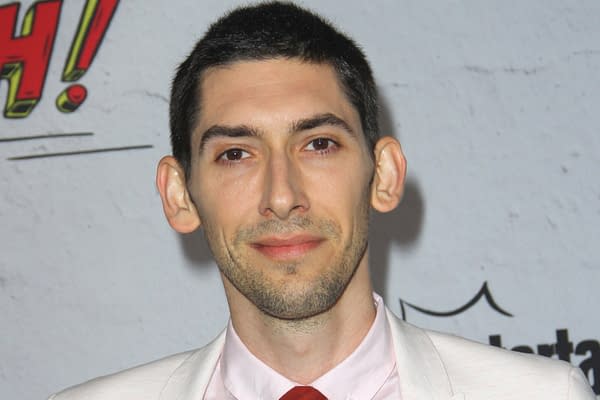 A man with short dark hair and a light suit smiles at the camera, standing in front of a textured background with bold text. He appears confident and is wearing a red tie.