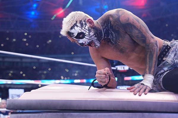 Darby Allin, painted in a dramatic black and white face design, kneels on a platform, appearing intense as he grips a weapon. The scene is set during a wrestling match at AEW All In in London, showcasing a high-adrenaline moment.