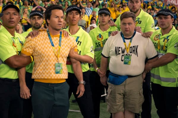 In a scene from 'Balls Up', Mark Wahlberg stands anxiously in a yellow patterned shirt, flanked by several uniformed security personnel, including Elijah Hauser who appears nervous while being escorted. The background features a cheering crowd in vibrant colors, emphasizing an intense moment.
