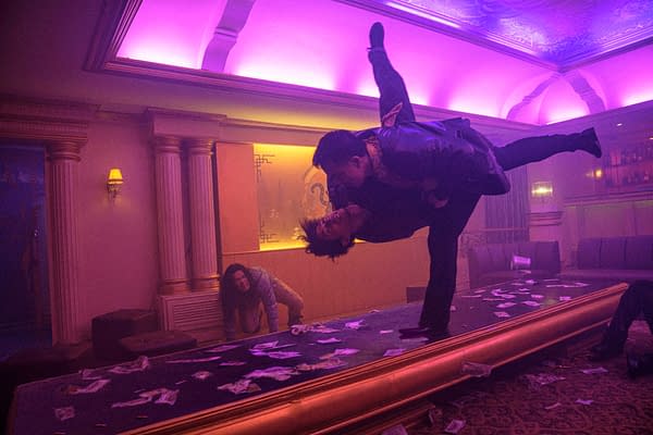 A dramatic action scene set in a dimly lit venue with purple lighting, featuring two men in a fierce struggle, one lifting the other above his head while a woman in the background appears distressed on the floor amid scattered cash.