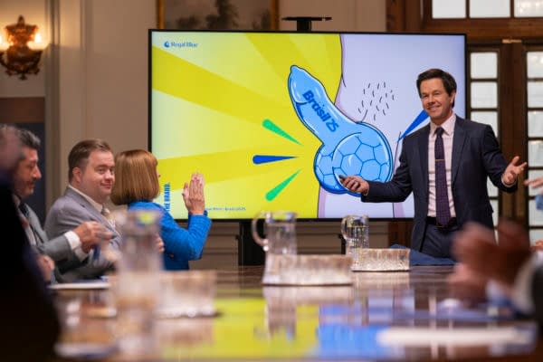In a conference room, a man in a suit enthusiastically presents an illustration featuring a large blue cartoon object resembling a soccer ball, alongside the text "Brasil" and bright yellow rays, while an audience of several people engages with him.