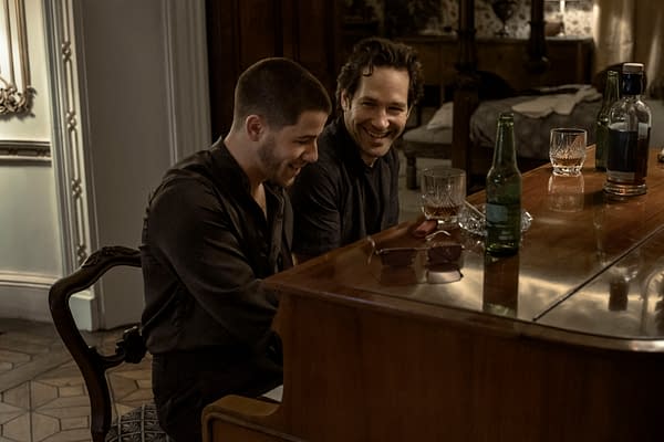 A scene from the film 'Power Ballad' featuring Nick Jonas as Danny and Paul Rudd as Rick. They sit together at a piano, sharing a moment of laughter, with drinks and glasses visible on the piano.