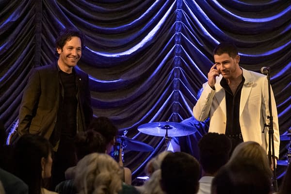 A scene from 'Power Ballad' featuring Paul Rudd and Nick Jonas, both smiling at an audience. The background is a dramatic curtain with blue lighting, and a drum set is visible behind them.
