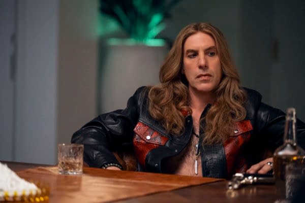 A man with long hair wearing a black leather jacket with red accents sits at a table, looking contemplative. A glass and a bottle are visible on the table along with a gun.