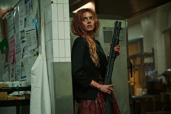 A scene from 'Ready Or Not 2: Here I Come' featuring a woman with long, messy hair holding a shotgun, standing against a wall in a kitchen environment. She appears tense and is wearing a black jacket over a bloodied dress.