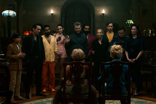 A dramatic scene from the film 'READY OR NOT 2: HERE I COME,' featuring a group of actors gathered in a dimly lit room. The characters appear engaged in a tense interaction, with one figure seated and facing others, creating an atmosphere of suspense.