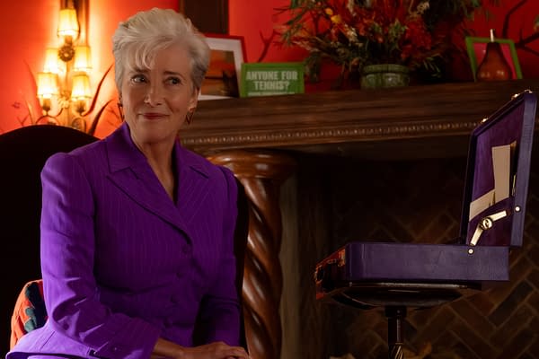 A scene from 'The Sheep Detectives' featuring Emma Thompson as Lydia Harbottle, wearing a bright purple suit and sitting beside an open purple case. The background is warmly lit with decorative plants and a fireplace, creating a cozy atmosphere.