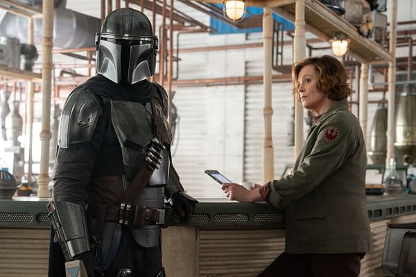 Mandalorian character in full armor stands next to Colonel Ward, who is seated at a bar-like counter, both appearing to engage in conversation. This scene is from Lucasfilm's 'The Mandalorian and Grogu.'