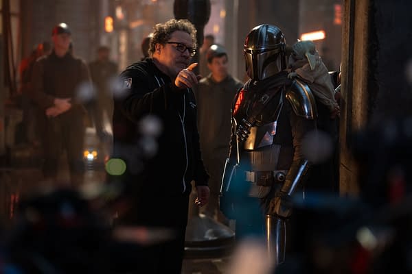 Behind-the-scenes image featuring director Jon Favreau discussing a scene with Pedro Pascal in full costume as the Mandalorian while Grogu is perched on his shoulder. The setting conveys a cinematic atmosphere from the production of 'The Mandalorian and Grogu' by Lucasfilm.
