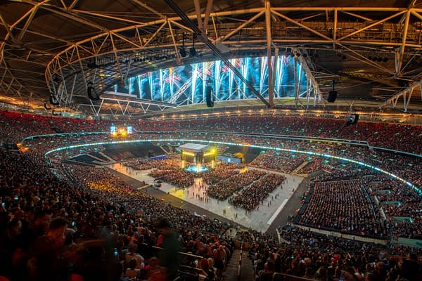 A wide shot of Wembley Stadium filled with a large crowd during the AEW All In event, showcasing bright lights and a stage set up for a live performance. The atmosphere is vibrant with fireworks lighting up the ceiling.