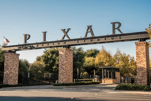 The entrance gate to Pixar Animation Studios, featuring large black lettering that spells "PIXAR" atop a brick structure, set against a clear blue sky and green trees.