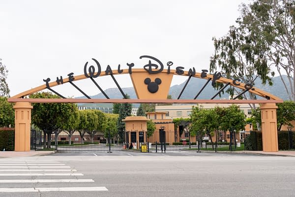 The iconic entrance gate of Walt Disney Studios in Burbank, California, prominently displays the company name and a Mickey Mouse silhouette, framed by greenery and a distant view of Hollywood hills.