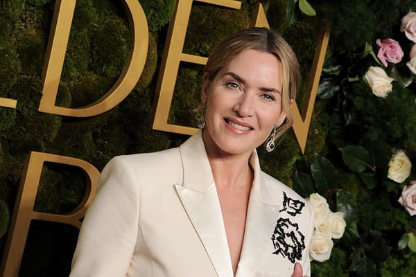 Kate Winslet smiles while posing at the Golden Globe Awards, wearing a stylish white outfit with intricate detailing. The background features greenery and soft rose accents, enhancing the elegant event atmosphere.