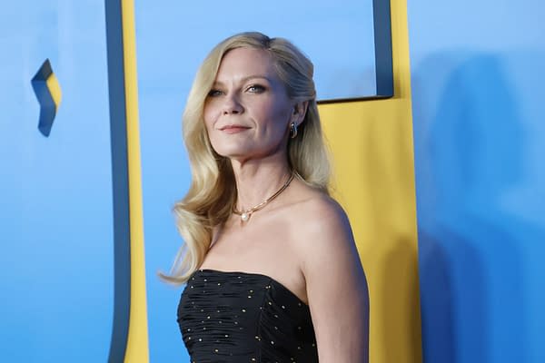 Kirsten Dunst poses elegantly in a black strapless dress adorned with sparkling accents, standing against a vibrant blue and yellow backdrop at a Hollywood premiere.