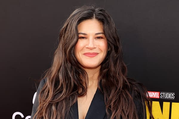 A woman with long, wavy dark hair smiles confidently, dressed in a black blazer, against a dark background with the Marvel Studios logo. This image is related to the announcement of new writers for the upcoming X-Men movie.
