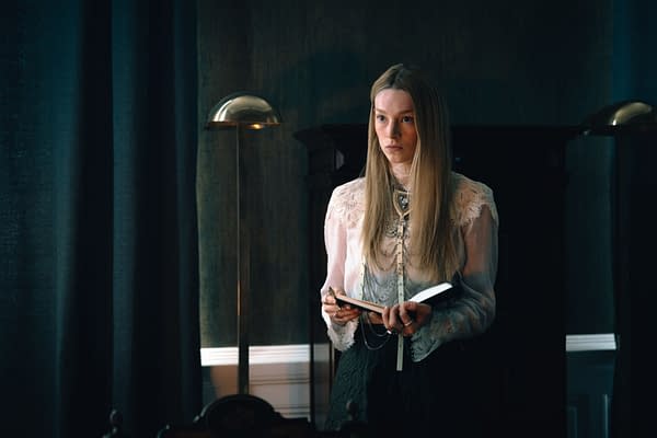 A woman with long blonde hair stands holding a book in a dimly lit room, wearing a vintage lace blouse and layered necklaces. She appears contemplative, set against a backdrop of dark curtains and a piece of furniture.