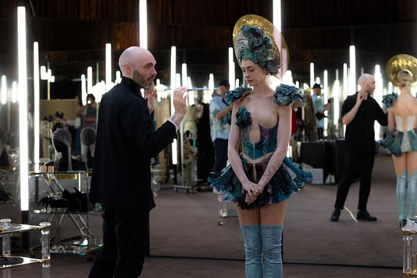 A fashion designer discusses details with a model in a striking, colorful outfit featuring feathers and bold patterns. The setting is a well-lit studio with mirrors reflecting various outfit designs.