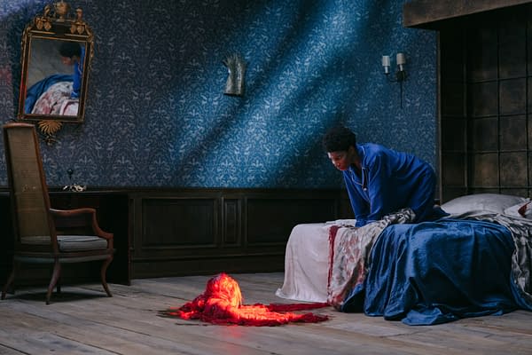A woman in a blue robe leans over a bed in a dimly lit room, gazing thoughtfully at a small, glowing red sculpture on the floor. The room features patterned wallpaper and antique furniture, creating a dramatic atmosphere.