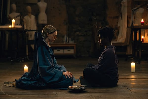 A scene depicting two women seated on a wooden floor, engaged in a serene moment. One woman wears a flowing blue garment, while the other is dressed in a dark top, with candles illuminating the space around them.