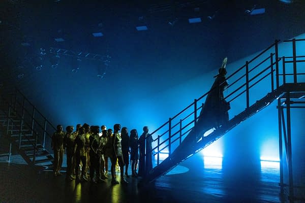 A performance scene featuring a figure resembling Mother Mary ascending a staircase, surrounded by a group of performers in golden costumes, under dramatic blue lighting.