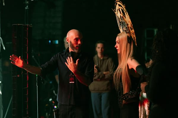 A behind-the-scenes moment featuring a director in a black shirt gesturing animatedly while speaking to a model wearing an elaborate gold headdress and a black outfit. The scene is illuminated by soft red lighting, with other crew members visible in the background.