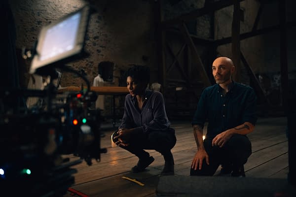 A film crew is captured in a vintage interior setting, featuring two individuals, one with curly hair in a purple shirt and another with a bald head in a dark button-up shirt, both focused on a camera setup. The atmosphere conveys an intimate and creative space, likely during a video production.