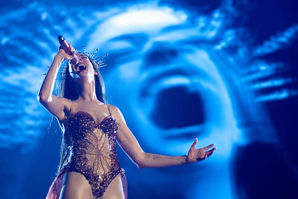A woman with long hair and a sparkling costume sings passionately into a microphone on stage, with a dramatic blue background featuring a shadowy figure of a screaming face.
